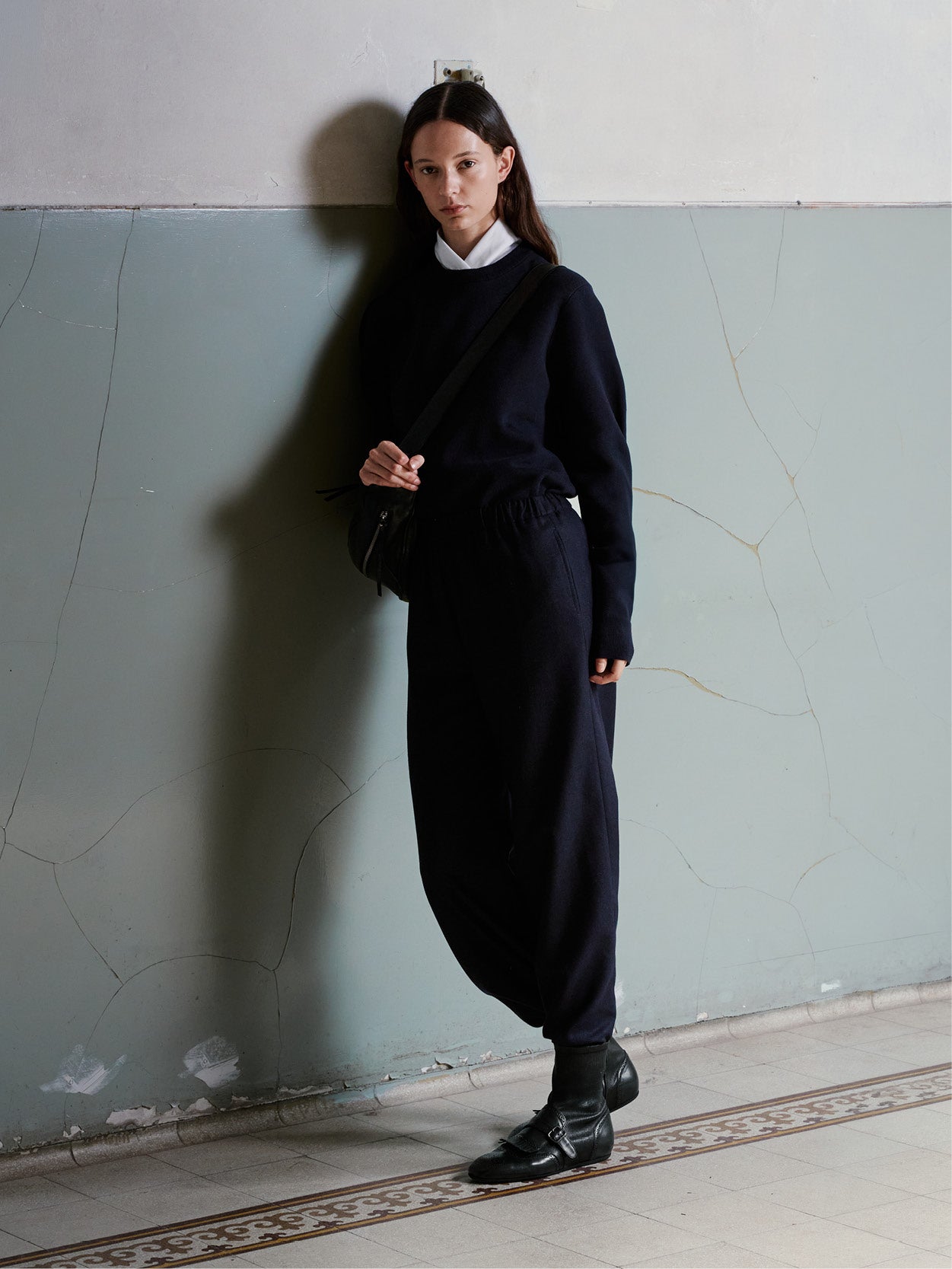 A model stands against a textured, cracked pale green wall wearing a dark navy long-sleeve knit top and matching loose-fitting navy pants, styled with a white collar, black shoes, and a black shoulder bag.