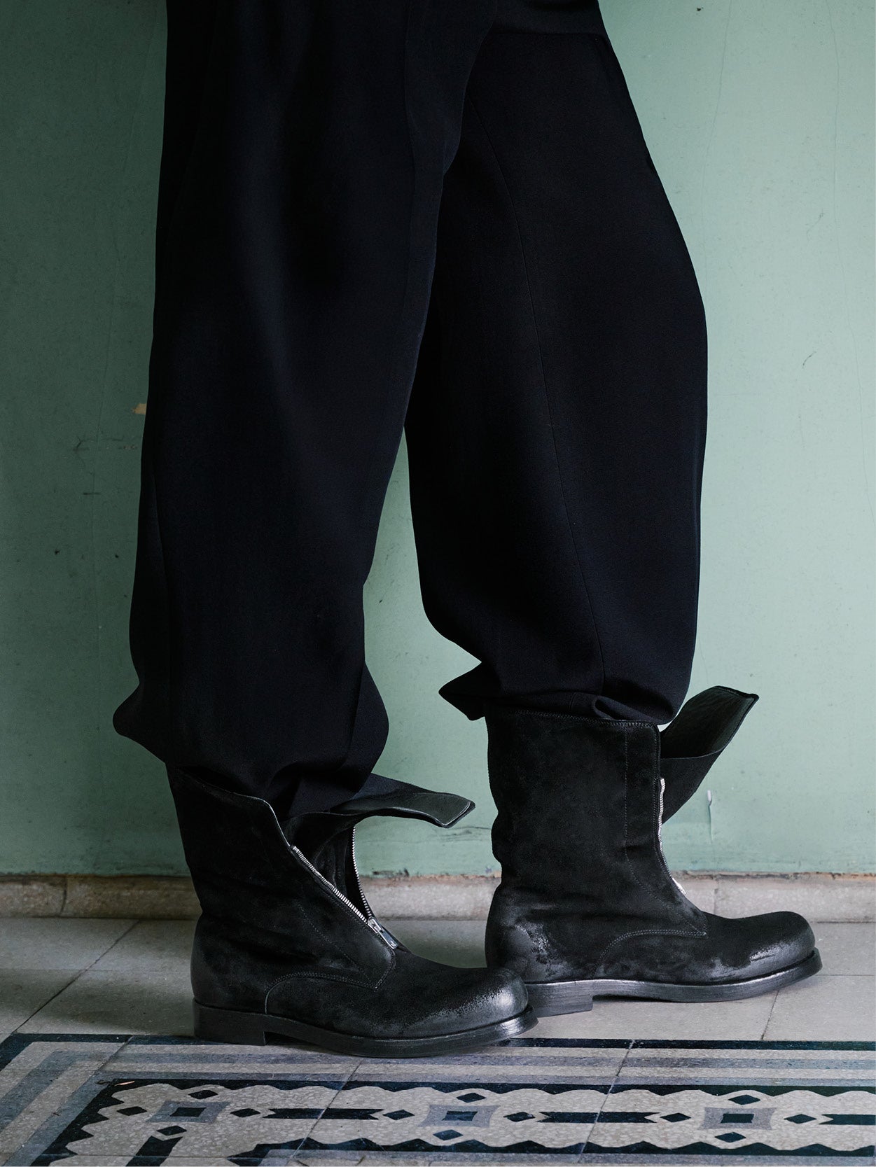 A person wearing loose black pants and black leather ankle boots with front zippers, standing indoors against a pale green wall.